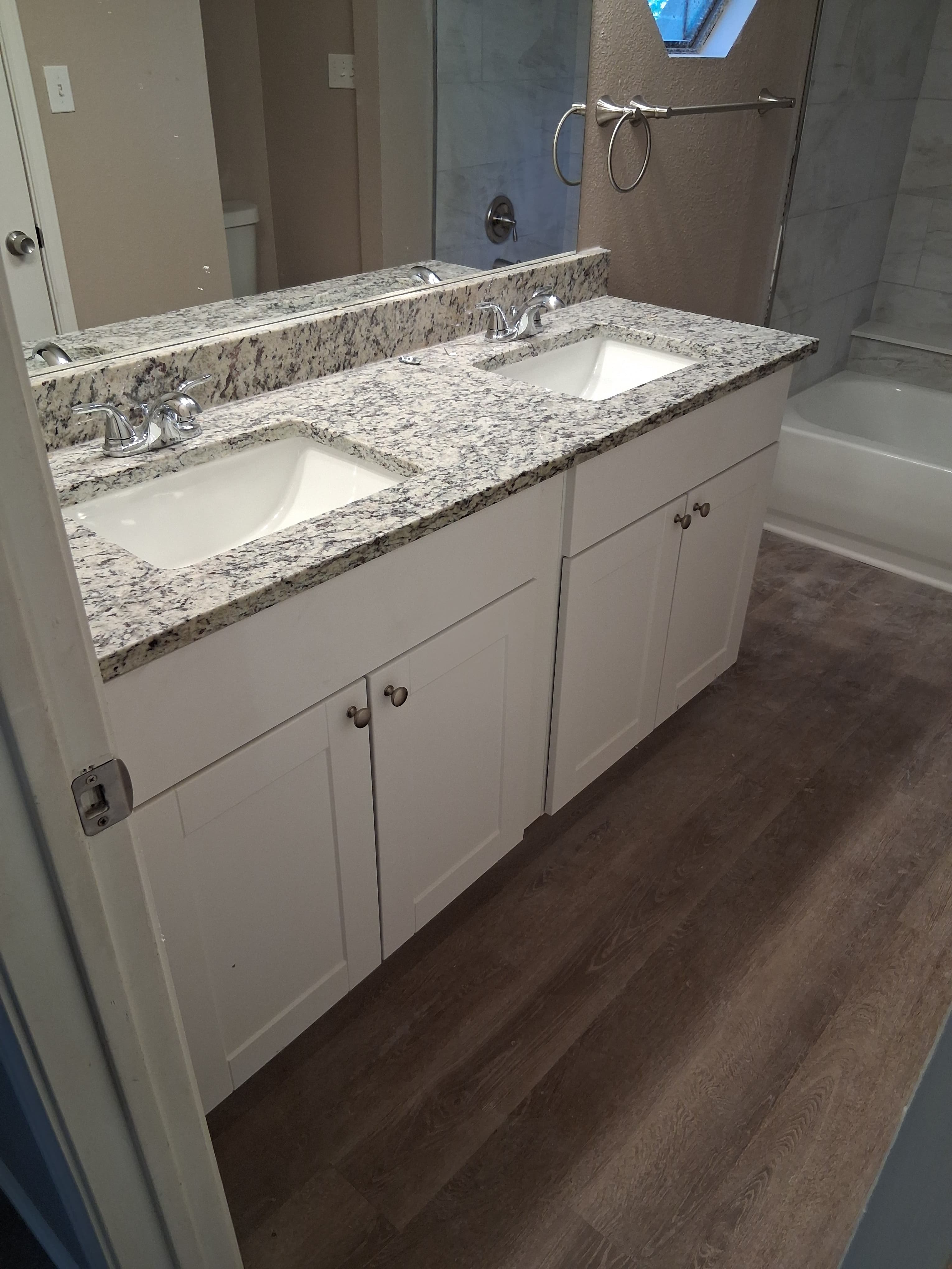 White double sink bathroom vanity with speckled granite countertops and dark wood-look flooring.