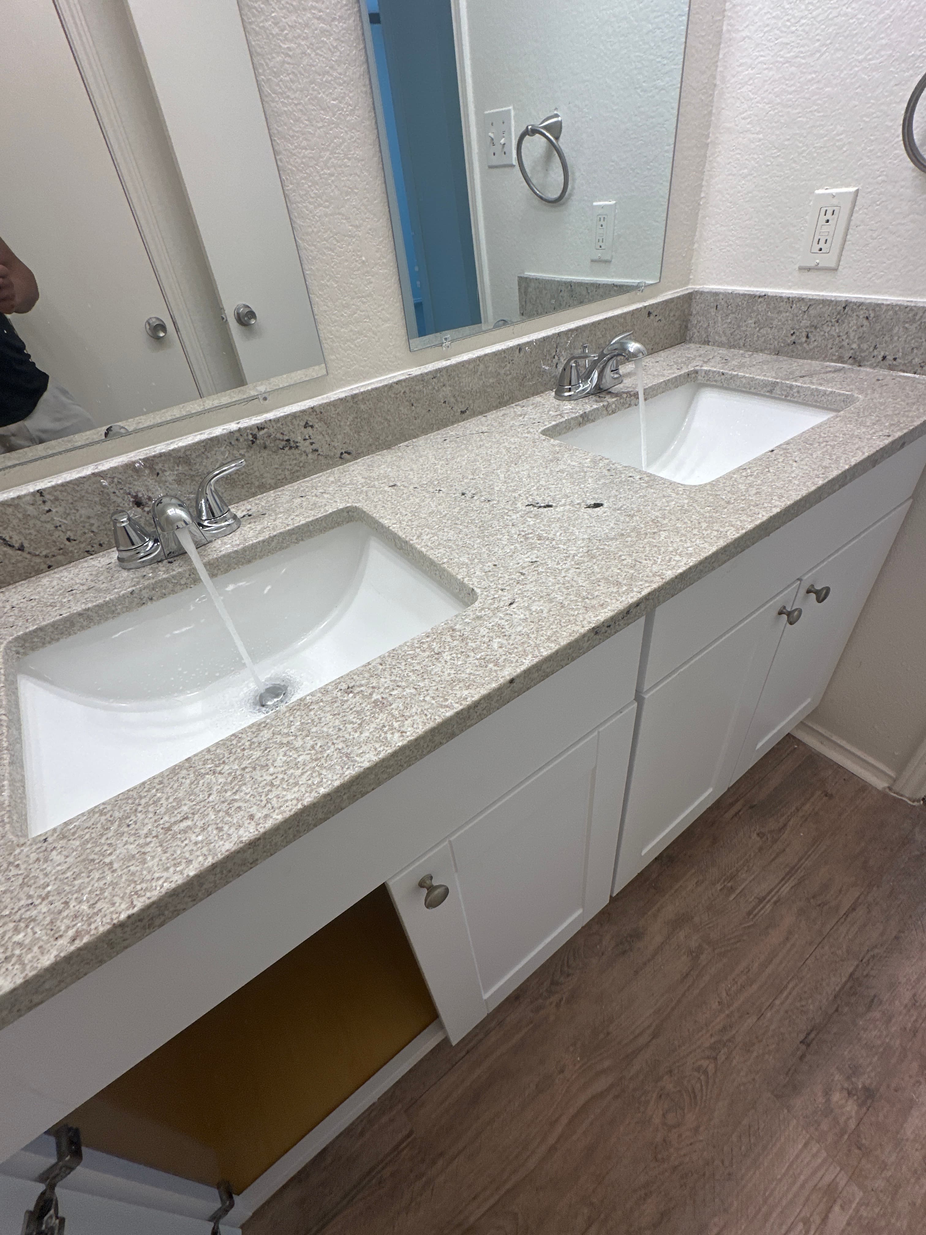 Double sink bathroom vanity with white cabinets, speckled countertops, and water running from faucets.