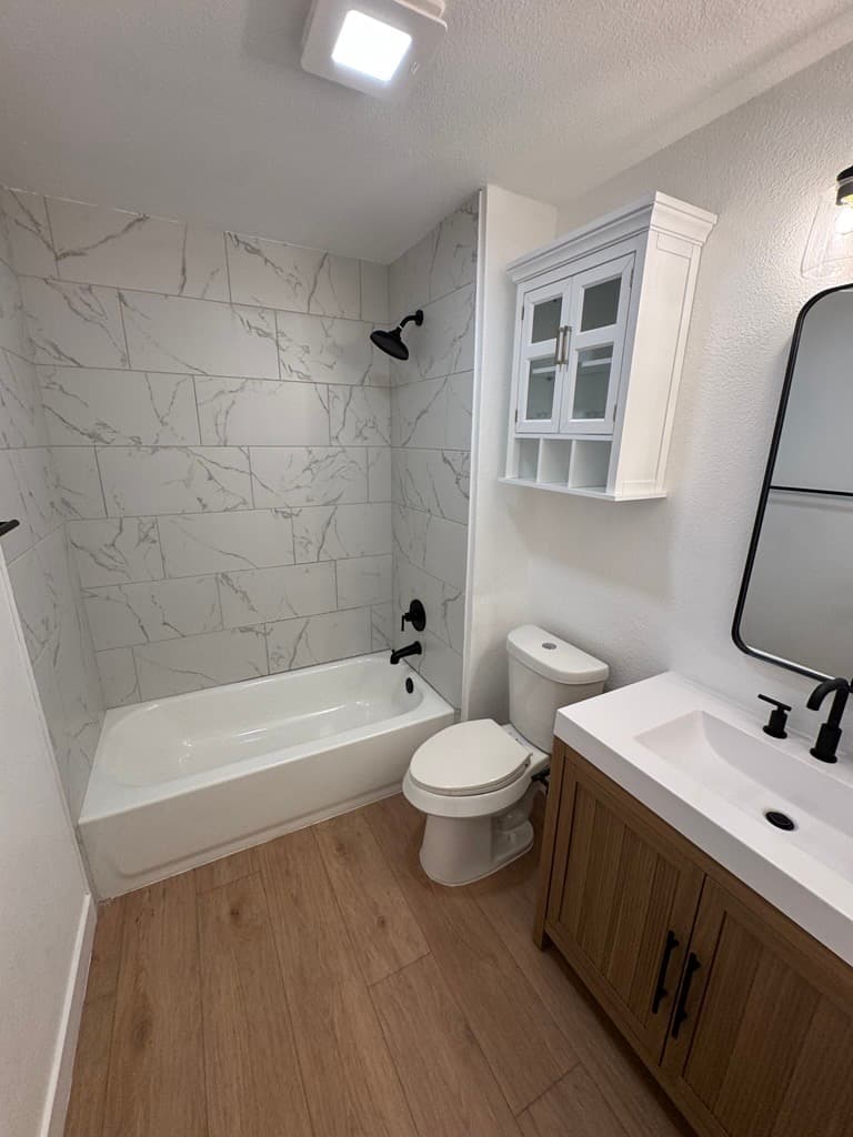 Modern bathroom with marble-patterned tile, white bathtub, toilet, and wooden vanity with black fixtures.