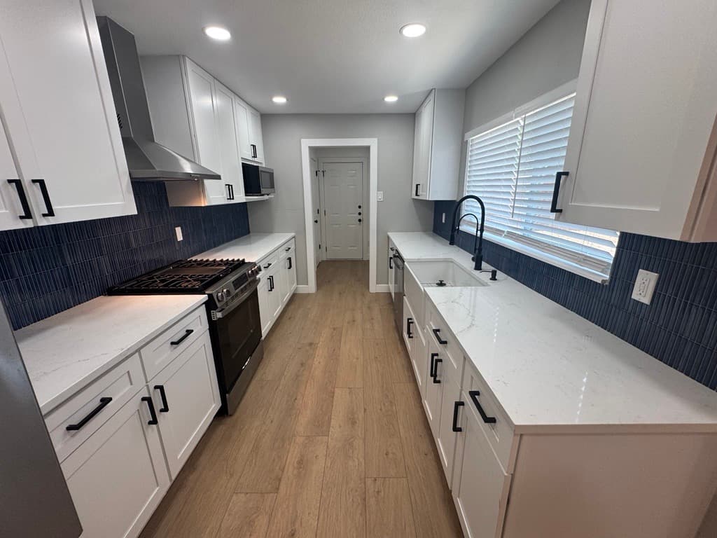 Modern galley kitchen featuring white cabinets, navy blue tile backsplash, and light wood flooring.