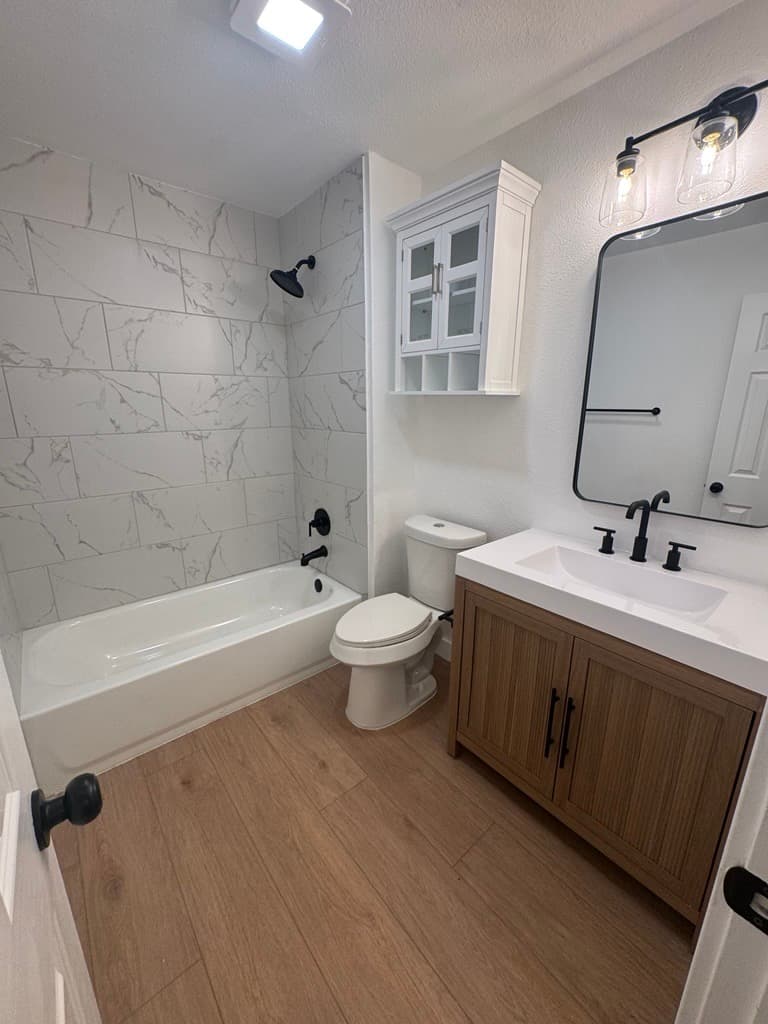 Modern bathroom with marble tile shower, wood vanity, black fixtures, and light wood floors.