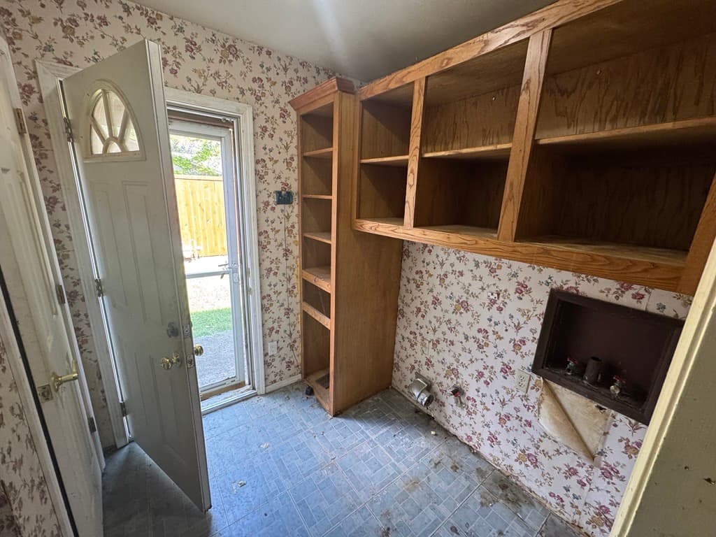 Small laundry room with floral wallpaper, wooden shelving, and an open door leading outdoors.