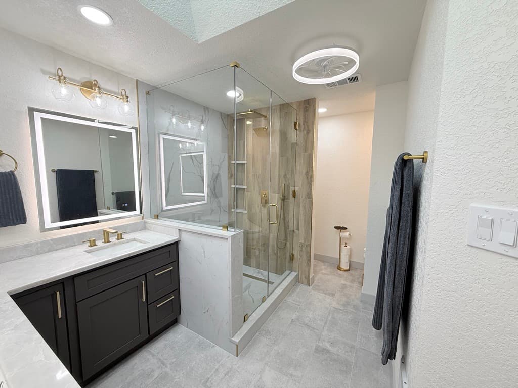 Modern bathroom with dark vanity, gold hardware, and glass shower with wood-look tile.