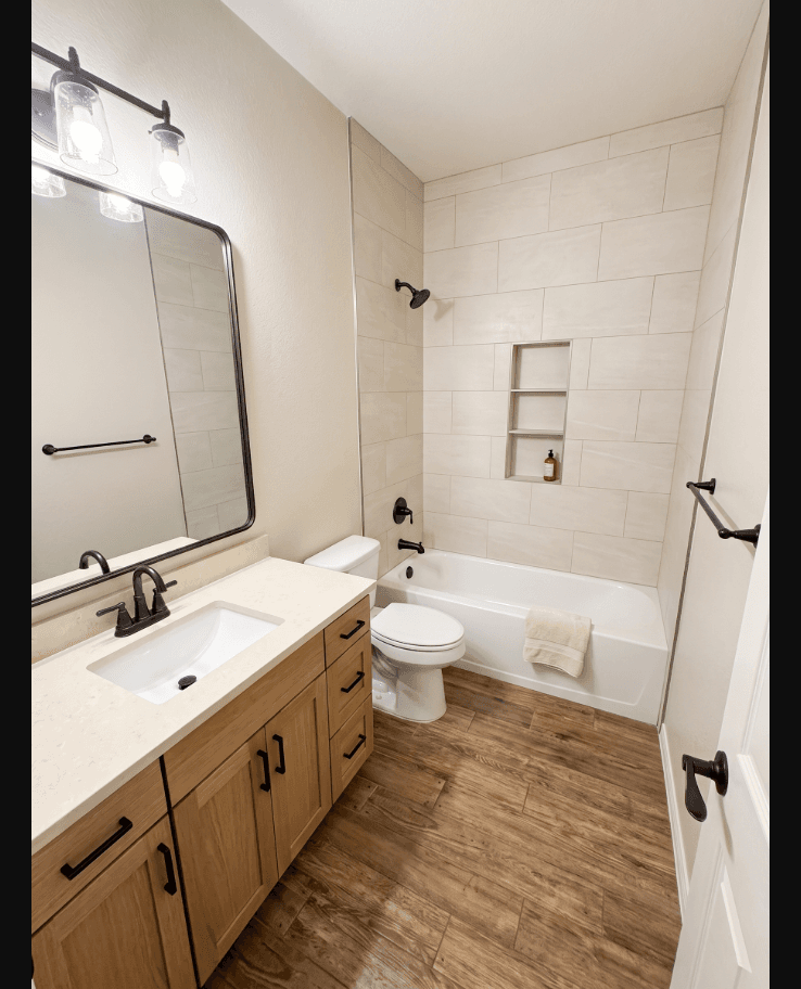 Modern bathroom featuring a light wood vanity, white bathtub with beige tile, and wood-look flooring.